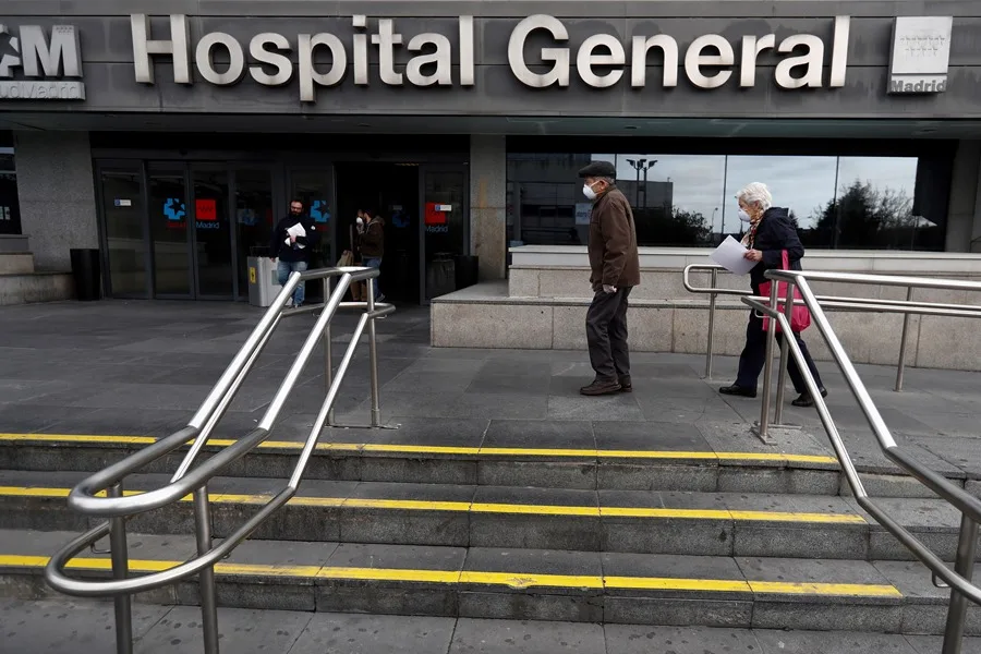 Dos personas mayores con mascarillas caminan junto a la entrada del hospital La Paz